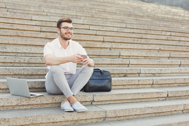 Un hombre de negocios sonriente con anteojos está enviando mensajes de texto a su teléfono mientras está sentado entre una computadora portátil y un maletín en las escaleras de piedra, copiando espacio. Concepto de tecnología, comunicación y negocio.