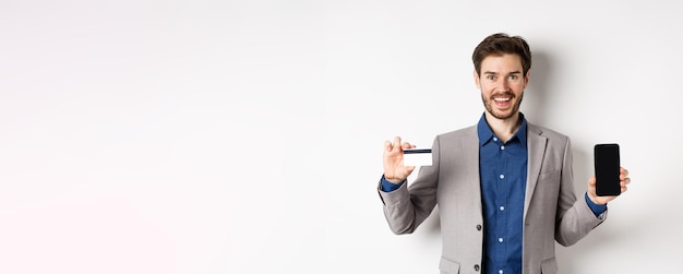 Foto hombre de negocios sonriente de compras en línea en traje que muestra una tarjeta de crédito de plástico con una pantalla de teléfono inteligente vacía