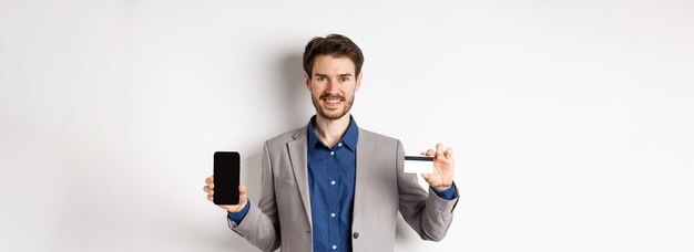 Hombre de negocios sonriente de compras en línea en traje que muestra una tarjeta de crédito de plástico con una pantalla de teléfono inteligente vacía