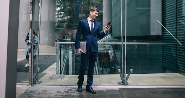 Hombre de negocios sonriente con elegante traje azul parado cerca de la pared de vidrio de un moderno edificio de oficinas con carpeta para documentos y teléfono inteligente en la mano