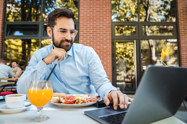 Un hombre de negocios sonriente está usando una computadora portátil está sentado en el restaurante al aire libre tomando un almuerzo y una bebida en un día soleado