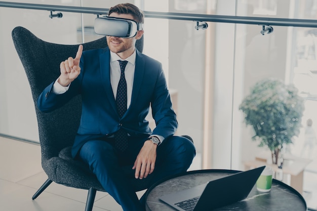 Hombre de negocios sonriente con gafas VR mientras está sentado en el vestíbulo de la oficina y trabaja en la computadora portátil