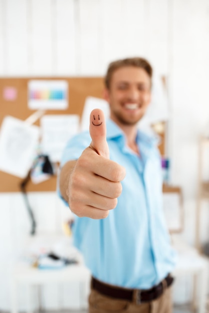 Foto hombre de negocios sonriente hermoso joven que muestra el pulgar para arriba con el dibujo de la cara divertida. centrarse en la mano.