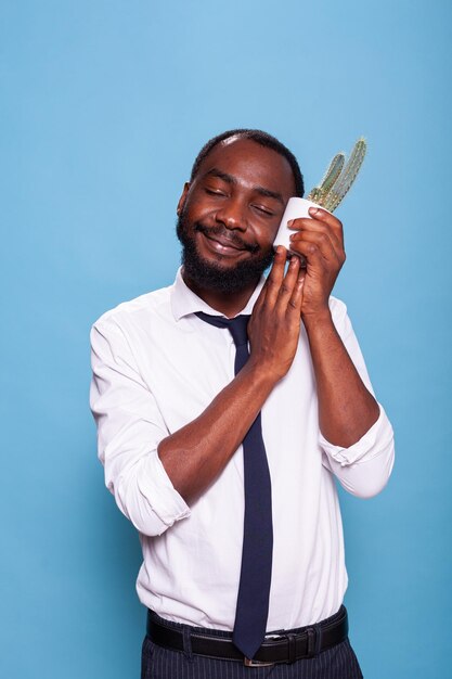 Hombre de negocios sonriente con los ojos cerrados presionando cactus en una olla blanca contra la mejilla mostrando amor por la planta de interior. Feliz oficinista afroamericano con personalidad cariñosa sosteniendo una planta en maceta.