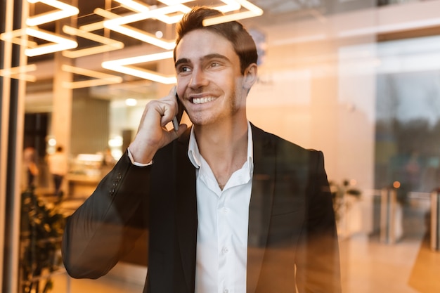 Hombre de negocios sonriente en traje hablando por teléfono detrás del vidrio y mirando a un lado