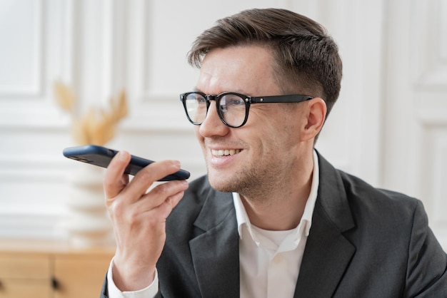 Un hombre de negocios sonriente en un traje usando comandos de voz en su teléfono en un espacio de oficina elegante y brillante
