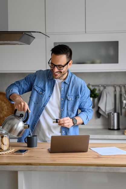 Un hombre de negocios sonriente vertiendo agua caliente en una taza de café mientras trabaja con una computadora portátil en casa
