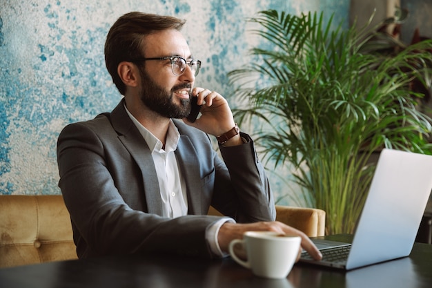 Hombre de negocios sonriente vistiendo traje hablando por teléfono móvil mientras está sentado en el café y trabajando en la computadora portátil