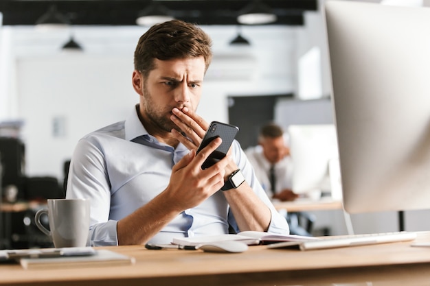 Hombre de negocios sorprendido con smartphone y tapándose la boca mientras está sentado junto a la mesa en la oficina