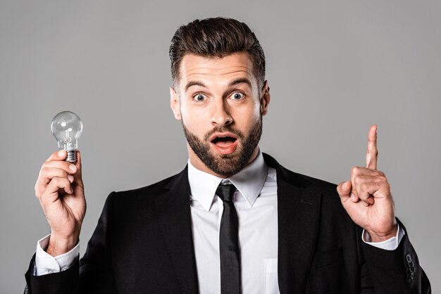 Foto hombre de negocios sorprendido en traje negro sosteniendo bombilla y mostrando gesto de idea aislado en gris