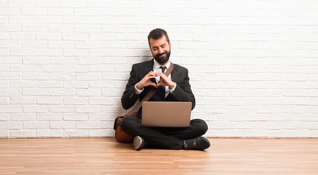 Hombre de negocios con su computadora portátil que se sienta en el piso que hace símbolo del corazón por las manos