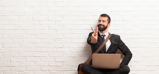 Hombre de negocios con su computadora portátil que se sienta en el suelo que sonríe y que muestra la muestra de la victoria