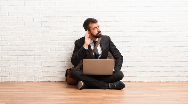 Hombre de negocios con su computadora portátil sentada en el piso escuchando algo poniendo la mano en la oreja