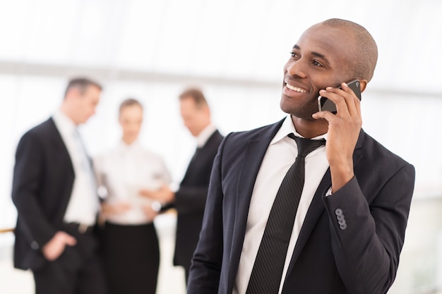 Hombre de negocios en el teléfono. Alegre joven africano en ropa formal hablando por teléfono móvil y sonriendo mientras sus colegas de pie en el fondo