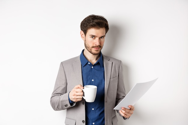 Hombre de negocios en traje leyendo documentos documentos y tomando café, sonriendo confiado a la cámara, fondo blanco.