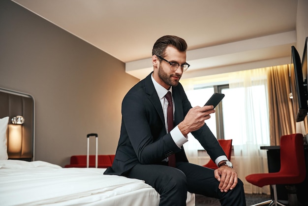 Foto hombre de negocios en traje de negocios sentado con el teléfono inteligente en la cama en la habitación del hotel