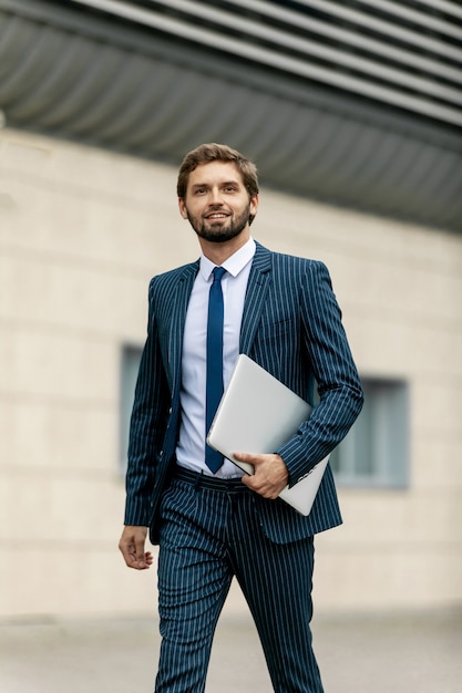 Hombre de negocios en traje con el portátil en la mano va a trabajar en el camino a la entrada de un edificio de oficinas de vidrio.