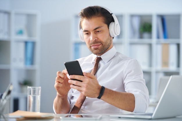 Hombre de negocios tranquilo contemporáneo en auriculares eligiendo la banda sonora de la lista de reproducción en el teléfono inteligente en el descanso