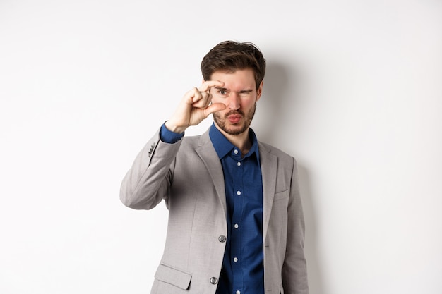 Hombre de negocios tratando de ver algo pequeño, mostrando el tamaño de una cosa pequeña con los dedos, de pie en traje sobre fondo blanco.