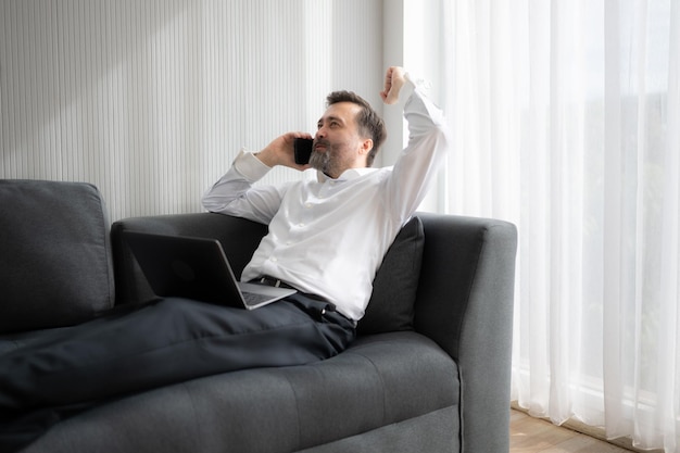 Hombre de negocios usando una computadora portátil y hablando por teléfono mientras está sentado en el sofá en casa