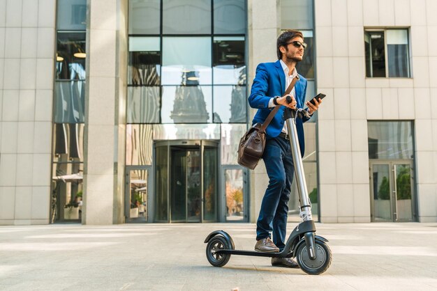 Hombre de negocios usando el teléfono celular mientras conduce un scooter eléctrico cerca del centro de negocios en la calle de la ciudad