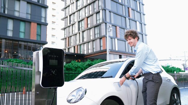 Hombre de negocios vestido con ambición progresiva apoyado en su vehículo eléctrico mientras está de pie en una estación de carga con un enchufe de cable de alimentación y un vehículo eléctrico alimentado por energía renovable