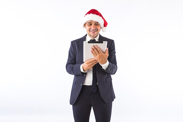 Hombre de negocios vestido con traje, corbata y sombrero de Navidad de Santa Claus, sonriendo, mirando su tableta, sobre fondo blanco. Concepto de celebración de Navidad.