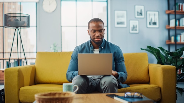 Hombre negro afroamericano guapo trabajando en una computadora portátil mientras está sentado en un sofá en una acogedora sala de estar Freelancer trabajando desde casa navegando por Internet usando redes sociales divirtiéndose en el piso