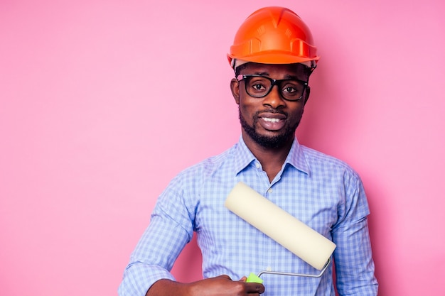Hombre negro afroamericano sosteniendo el rodillo de pintura en la mano pinta la pared en color rosa .Feliz constructor africano pintando dentro de la casa, el empresario usa un casco casco.