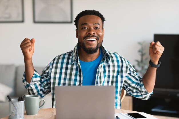 Hombre negro alegre usando computadora portátil gesticulando sí en casa