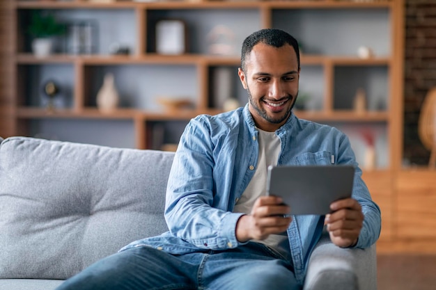 Hombre negro alegre viendo videos en una tableta digital mientras se relaja en el sofá