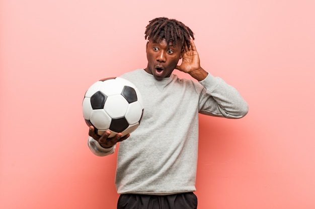 Hombre negro de la aptitud joven que sostiene un balón de fútbol que intenta escuchar un chisme.