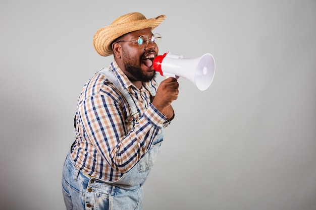 Hombre negro brasileño vestido con ropa de campo de festa junina festa de sao joao arraia usando megáfono gritando promoción anunciando descuento publicitario