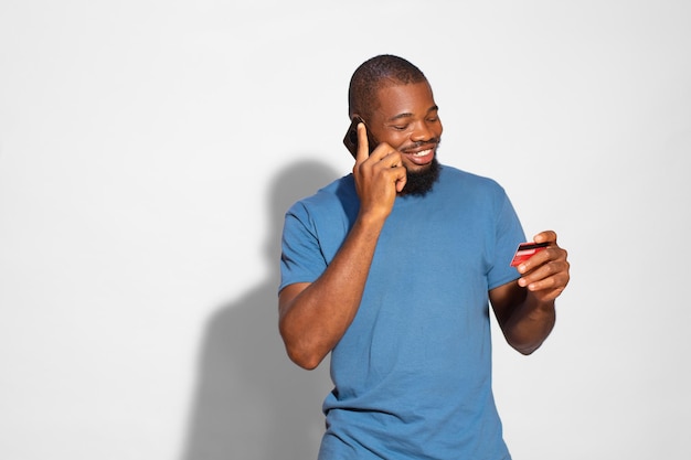 Hombre negro con una camiseta azul sonriendo y hablando por teléfono mirando una tarjeta bancaria en la mano