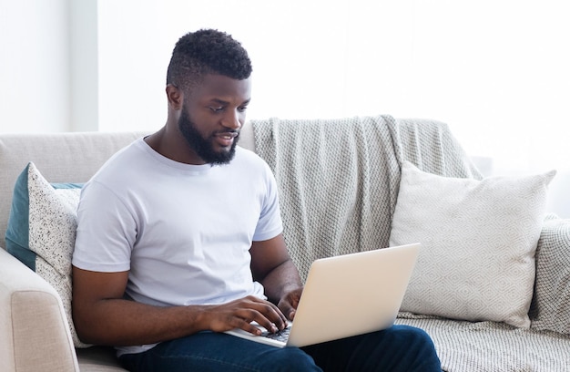 Un hombre negro con una camiseta blanca se sienta en un sofá en una sala de estar escribiendo en una computadora portátil que tiene