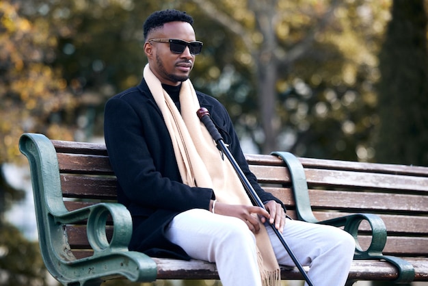 Un hombre negro ciego estaciona y se sienta con gafas de sol, bastón y se relaja junto a los árboles, paz y pensamiento. Chico africano, moda vintage y joven con gafas para discapacitados y viajes en la naturaleza.