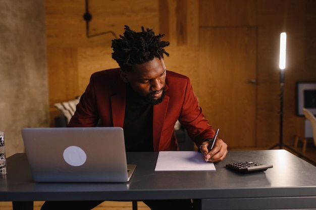 Un hombre negro concentrado en un traje está trabajando en una computadora portátil, trabajo remoto desde casa, foto de alta calidad