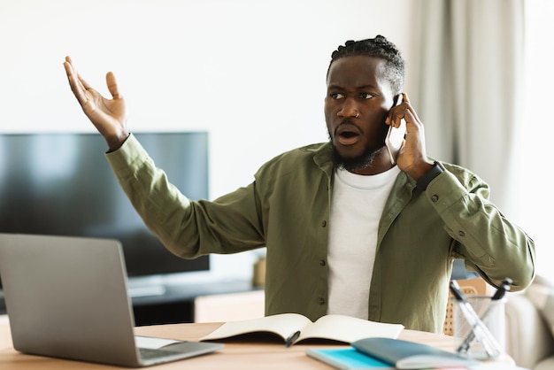 Hombre negro descontento hablando por teléfono celular y usando pc que tiene problemas con la conexión a internet en casa