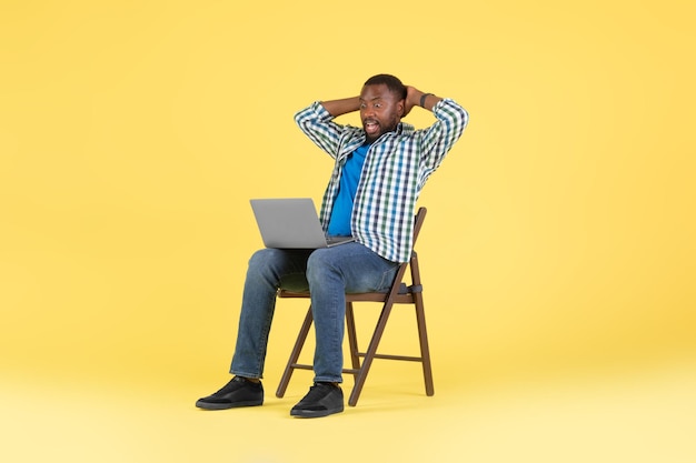 Hombre negro emocional mirando portátil sentado sobre fondo amarillo