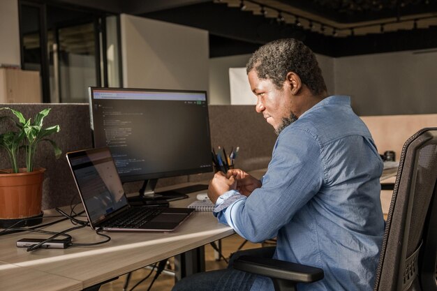 Hombre negro escribiendo líneas de código en una PC de escritorio con dos monitores y una computadora portátil a un lado en una oficina elegante