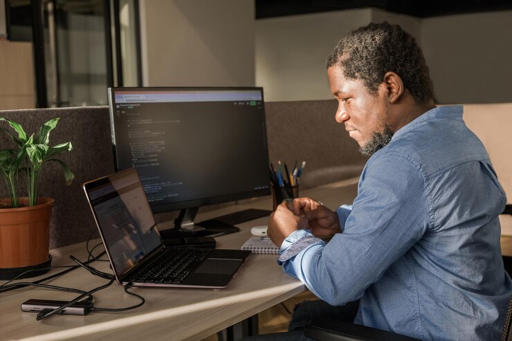 Foto hombre negro escribiendo líneas de código en una pc de escritorio con dos monitores y una computadora portátil a un lado en una oficina elegante