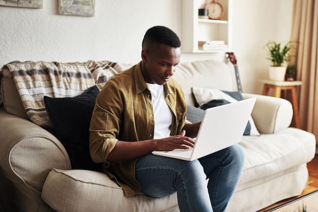 Hombre negro escribiendo y sofá con portátil para comunicación correo electrónico o navegación en línea en casa Joven hombre africano o freelance en tecnología para red de trabajo remoto o conexión a Internet