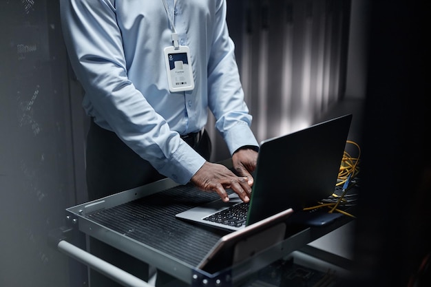 Hombre negro escribiendo en el teclado de la computadora portátil mientras configura la red en la sala de servidores