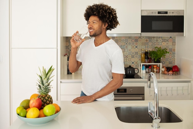 Hombre negro feliz bebiendo agua en medio de la preparación de alimentos en la cocina