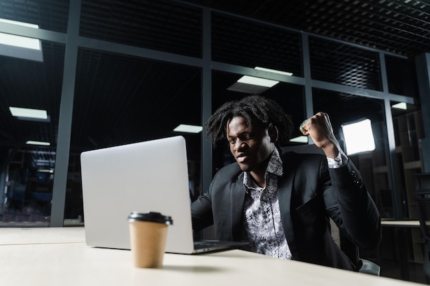 El hombre negro feliz con los puños apretados está mirando la pantalla de la computadora portátil y se regocija de terminar su trabajo.