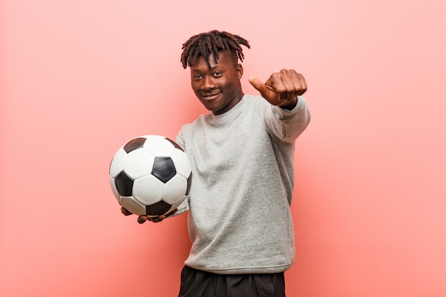 Hombre negro fitness joven sosteniendo un balón de fútbol sonrisas alegres apuntando al frente.