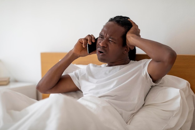 Hombre negro frustrado hablando por teléfono celular mientras está sentado en la cama en casa