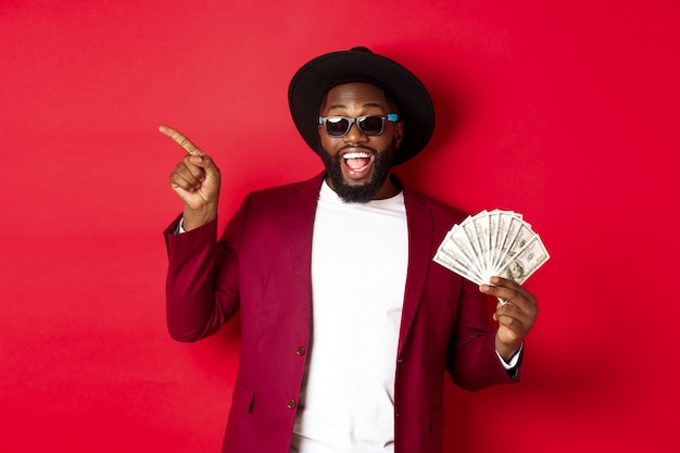 Hombre negro guapo y elegante apuntando con el dedo hacia la izquierda mientras muestra dinero, sosteniendo dólares y mostrando el logotipo, de pie sobre fondo rojo