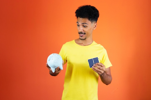 Hombre negro guapo sosteniendo una alcancía y un panel solar aislado en naranja Hombre en una camisa amarilla aislada