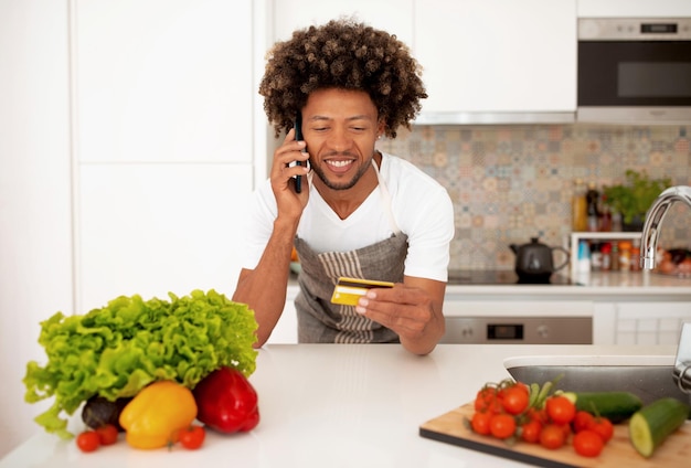 Hombre negro hablando por teléfono con tarjeta de crédito en la cocina